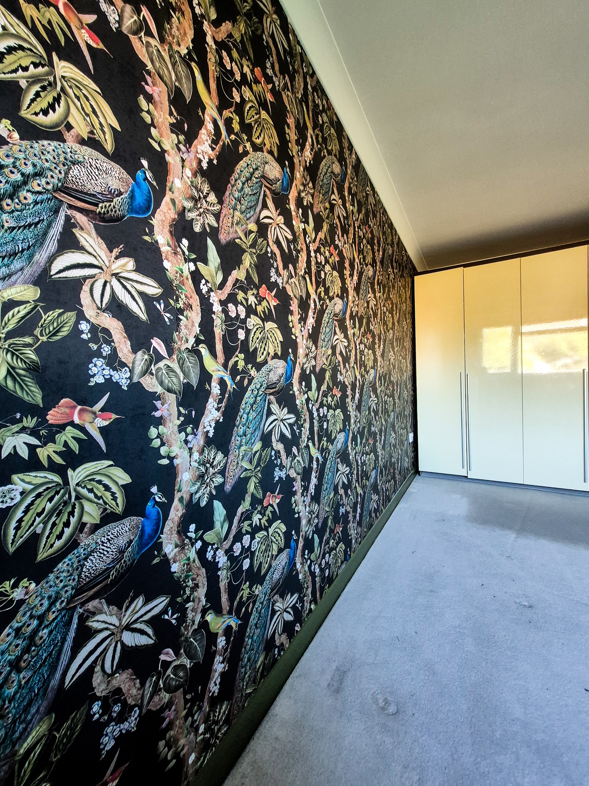 Peacock and botanical feature wallpaper along a bedroom wall beside cream wardrobes